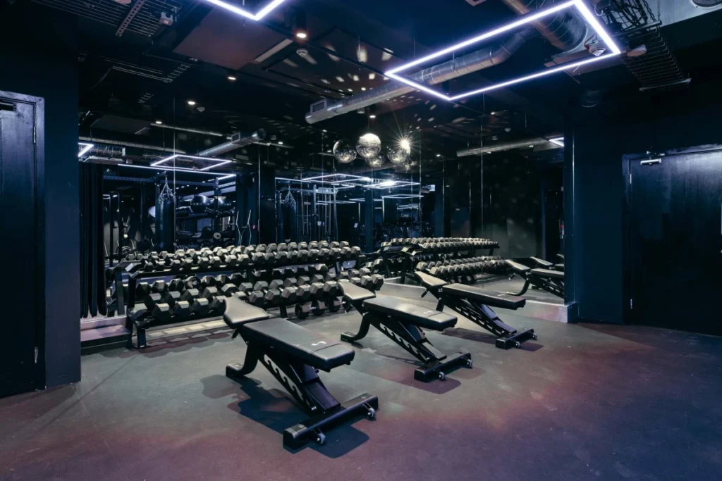 Adjustable weight benches and dumbbell racks beneath geometric neon lighting and disco balls