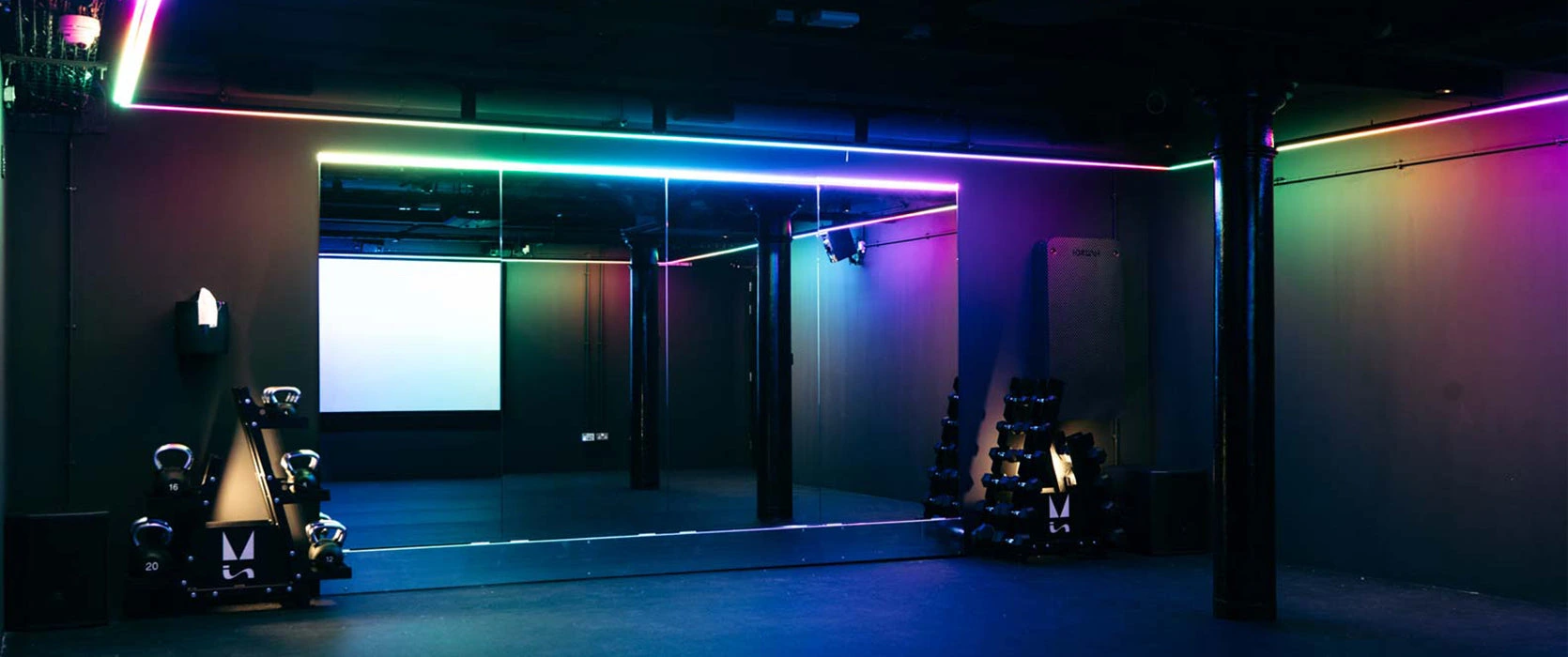 Mirrored studio space with colourful RGB lighting, kettlebell rack, and projector screen