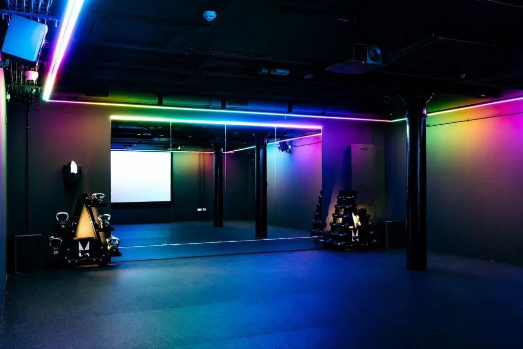 Mirrored studio space with colourful RGB lighting, kettlebell rack, and projector screen